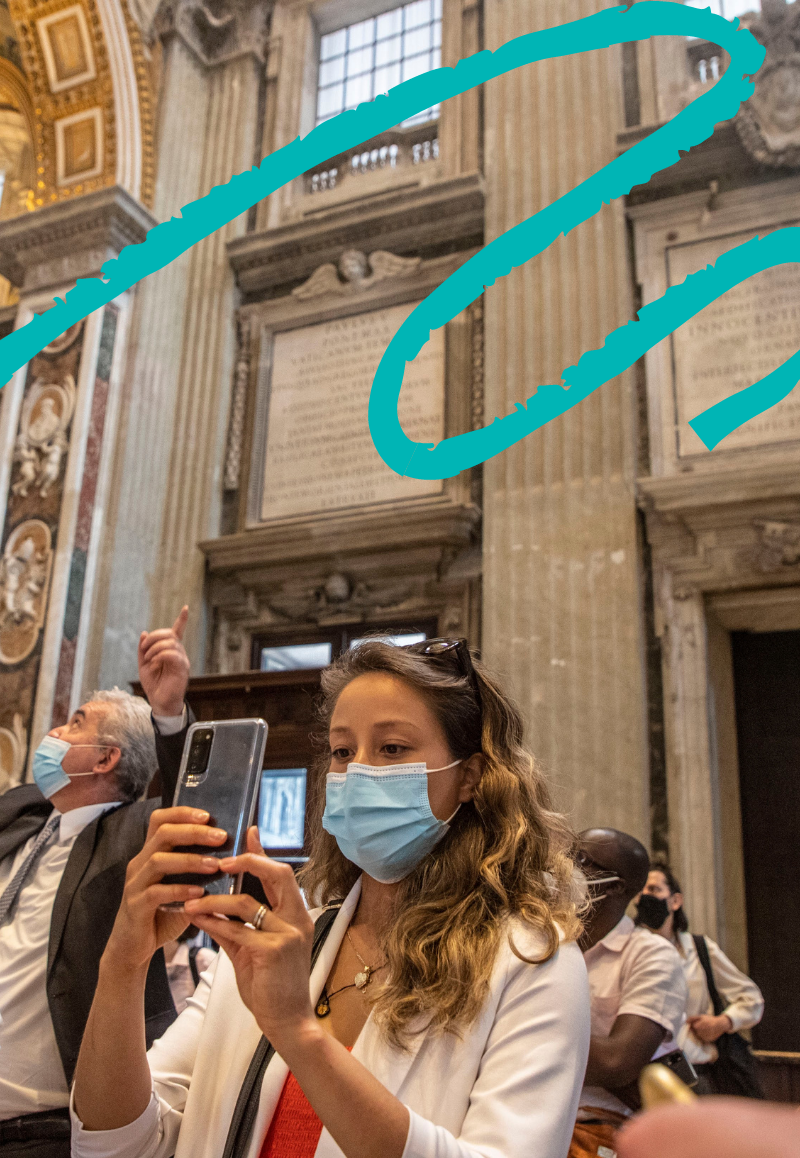 A group of people standing in a church-like building, the image focuses on a brunette Caucasian woman wearing a face mask and holding a phone.