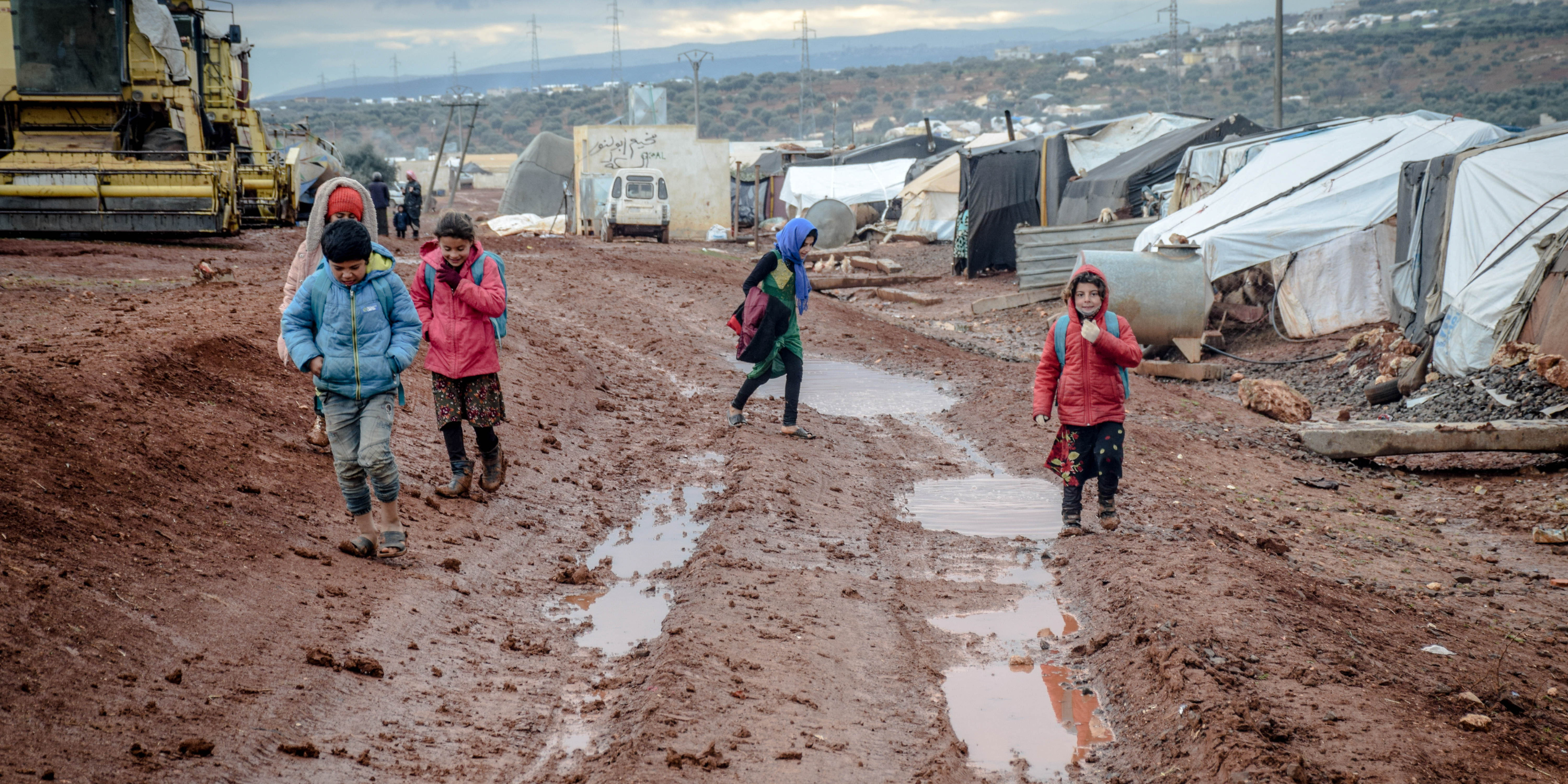Children walking in muddy field, along shelters at refugee camp
