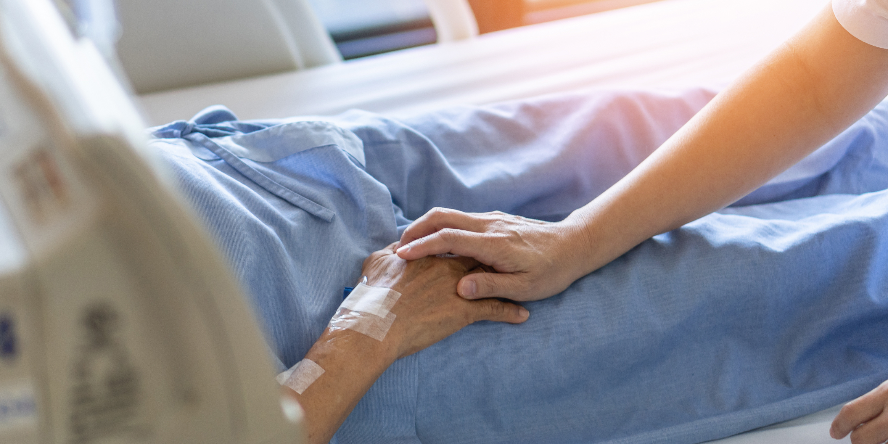 A hand resting on another hand of a person who is laying in a hospital bed, connected to IVs.