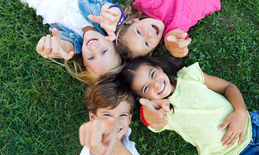 A birds eye view of a group of children laying down on the grass, smiling and pointing at the camera.