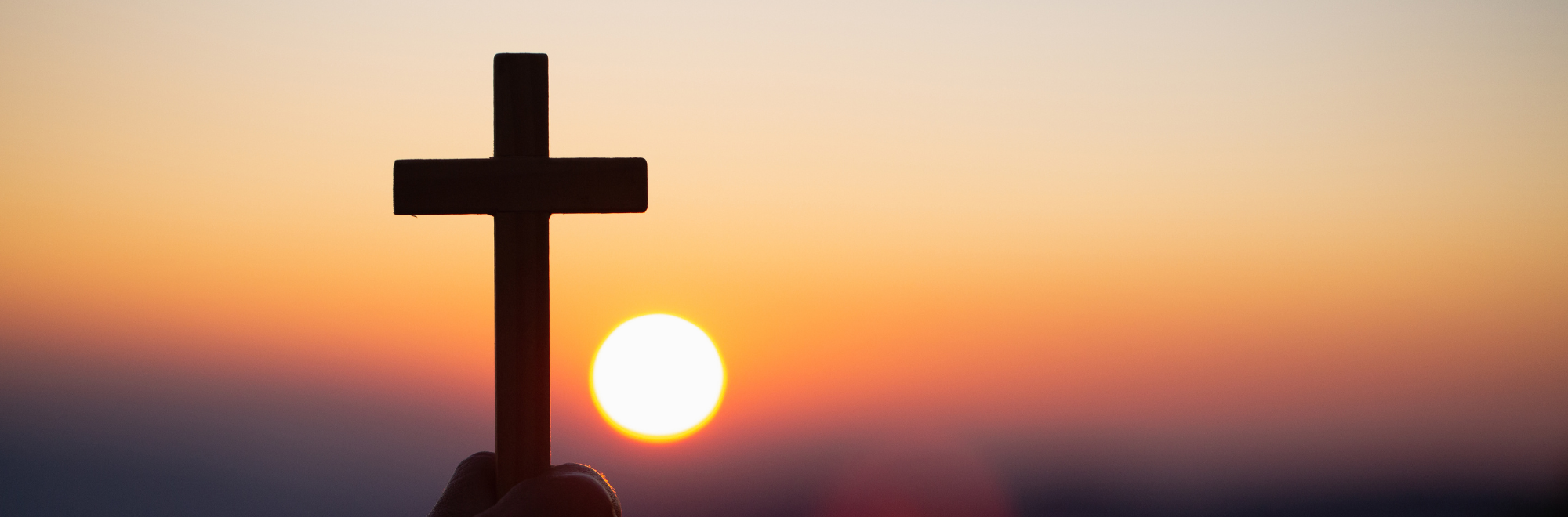 A close-up of someone holding a wooden cross, with a faded sunset in the background.